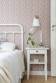 Flower in vase and brass candlestick on bedside table, crocheted blanket on metal bed in bedroom with floral wallpaper