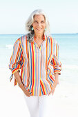 A mature woman with white hair on a beach wearing a striped shirt and white summer trousers