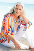 A mature woman with white hair on a beach wearing a striped shirt and white summer trousers