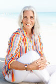 A mature woman with white hair on a beach wearing a striped shirt and white summer trousers