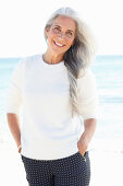 Reife Frau mit weißen Haaren in weißem Pullover und gepunkteter Hose am Strand