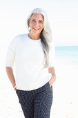 A mature woman with white hair on a beach wearing a white jumper and polka dot trousers