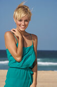A blonde woman with short hair on a beach wearing a turquoise dress