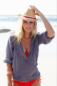 A blonde woman on a beach wearing a hat, a red bikini and a blue blouse