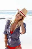 Blonde Frau mit Hut in rotem Bikini und blauer Bluse am Strand