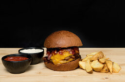 Hamburger mit Kartoffelspalten und zweierlei Dips vor schwarzem Hintergrund