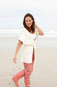 A brunette woman wearing a short-sleeved cardigan, pink trousers and wellie boots on a beach