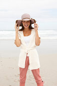 Brünette Frau mit Hut in Strickjacke mit kurzen Ärmeln und rosa Hose am Meer