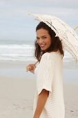 A brunette woman by the sea wearing a short-sleeved cardigan and holding a parasol