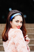 A young brunette woman wearing a pink woollen coat and a blue hairband
