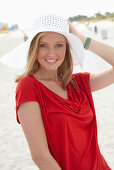 A blonde woman on a beach wearing a red top and a white hat