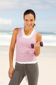 A young brunette woman by the sea wearing sports clothes with a dumbbell