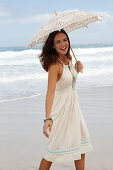 A brunette woman by the sea wearing a white summer dress and holding a parasol