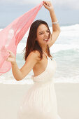 A brunette woman by the sea wearing a white summer dress and holding a pink shawl