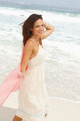 A brunette woman by the sea wearing a white summer dress and holding a pink shawl