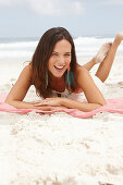 A brunette woman lying on a pink shawl by the sea wearing a white summer dress