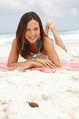 A brunette woman lying on a pink shawl by the sea wearing a white summer dress