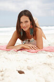 A brunette woman lying on a pink shawl by the sea wearing a white summer dress