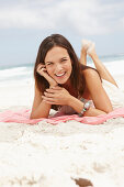 A brunette woman lying on a pink shawl by the sea wearing a white summer dress