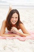 A brunette woman lying on a pink shawl by the sea wearing a white summer dress