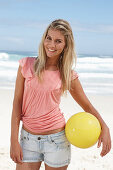 A young woman on a beach with a yellow ball wearing a pink top and denim shorts