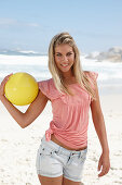 Junge Frau mit gelbem Ball im rosa Top und Jeansshorts am Strand