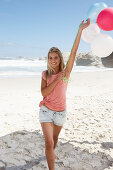 A young woman on a beach with balloons wearing a pink top and denim shorts