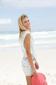 A blonde woman on the beach wearing a light t-shirt and denim shorts holding a red hat