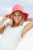 A blonde woman on the beach wearing a light t-shirt and a red hat