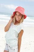 A blonde woman on the beach wearing a light t-shirt and denim shorts holding a red hat