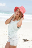 A blonde woman on the beach wearing a light t-shirt and denim shorts holding a red hat