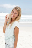 A blonde woman on the beach wearing a light t-shirt and holding a shell