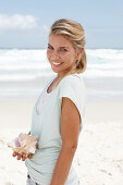 A blonde woman on the beach wearing a light t-shirt and holding a shell