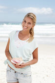 A blonde woman on the beach wearing a light t-shirt and holding a shell