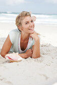 Blonde Frau in hellem T-Shirt am Strand liegend, vor ihr Muschelschale