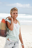 A blonde woman on the beach wearing a light t-shirt and denim shorts holding a wicker bag