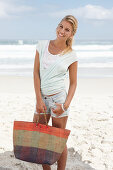 A blonde woman on the beach wearing a light t-shirt and denim shorts holding a wicker bag