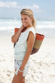 A blonde woman on the beach wearing a light t-shirt and denim shorts holding a wicker bag