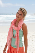 A blonde woman on a beach wearing a scarf, a turquoise top and a salmon pink skirt