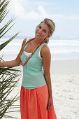 Blonde Frau mit Halskette in türkisgrünem Top und lachsfarbenem Rock am Strand