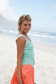 A blonde woman on a beach wearing a turquoise top and a salmon-coloured skirt