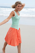 A blonde woman on a beach wearing a turquoise top and a salmon-coloured skirt