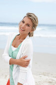 A blonde woman on a beach wearing a turquoise top and a white cardigan