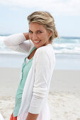 A blonde woman on a beach wearing a turquoise top and a white cardigan