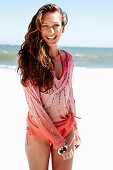 A brunette woman on a beach wearing a pink sequinned blouse and a bikini