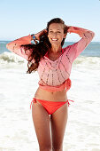 A brunette woman on a beach wearing a pink sequinned blouse and a bikini