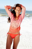 A brunette woman on a beach wearing a pink sequinned blouse and a bikini