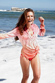 A brunette woman on a beach wearing a pink sequinned blouse and a bikini