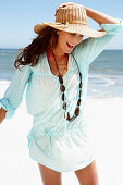 A brunette woman wearing a hat, a necklace and a light-blue beach dress by the sea