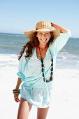 A brunette woman wearing a hat, a necklace and a light-blue beach dress by the sea